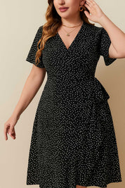Plus-size black polka-dot wrap mini dress with V-neck and side tie waist, soft A-line sway — front three-quarter view