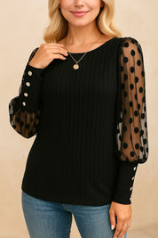 Black rib-knit blouse with sheer polka-dot sleeves and button cuffs, clean round neck — left three-quarter view