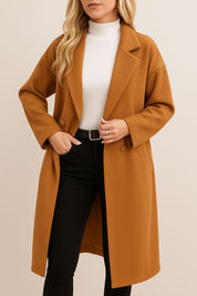 Front view of the caramel longline coat showing the notched lapel, covered buttons and a hand in the slant welt pocket; drop-shoulder cut.