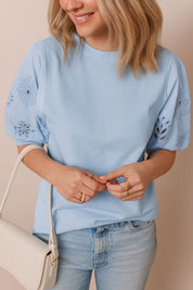 Close front detail view of a light blue puff-sleeve blouse with floral eyelet embroidery on the sleeves and a clean crew neckline
