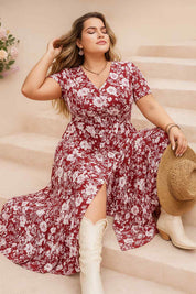 Confident woman in a red floral plus-size maxi dress with V-neck and side slit, sitting gracefully with white boots and holding a straw hat — boho cowgirl vibe.
