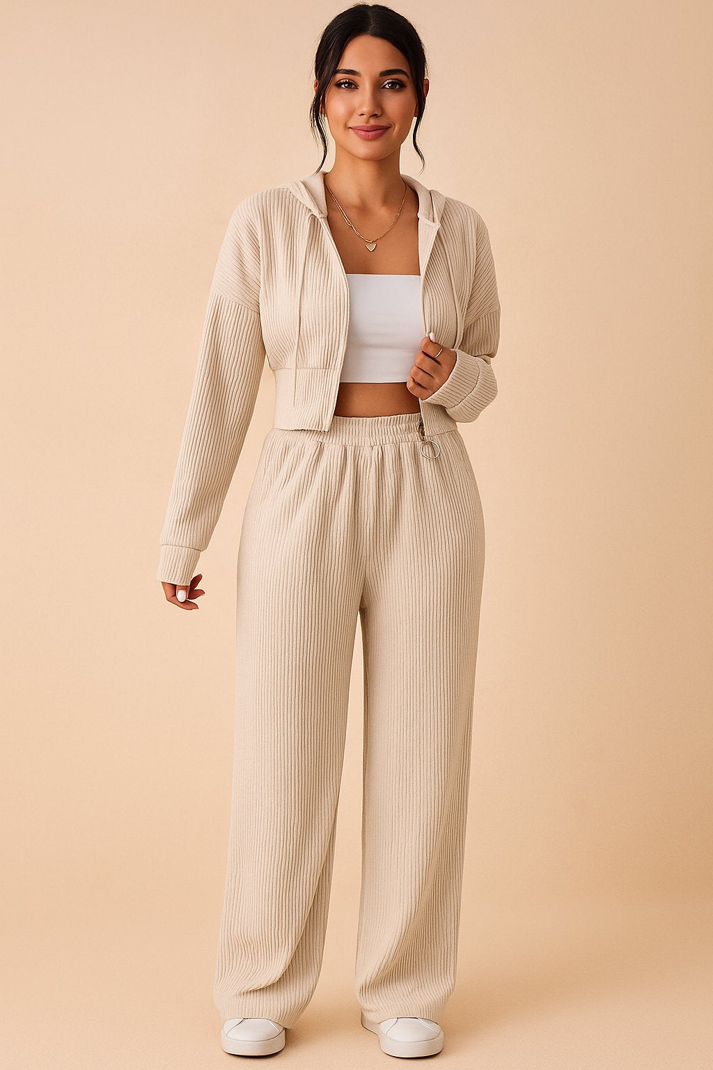 Pastel beige ribbed jumpsuit featuring zip‑up hoodie and elastic waistband, model kneeling.