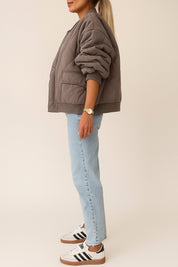 “Left side view of the taupe quilted bomber jacket with rib-knit hem and cuffs, wave quilting, and a snap-flap pocket at the hip