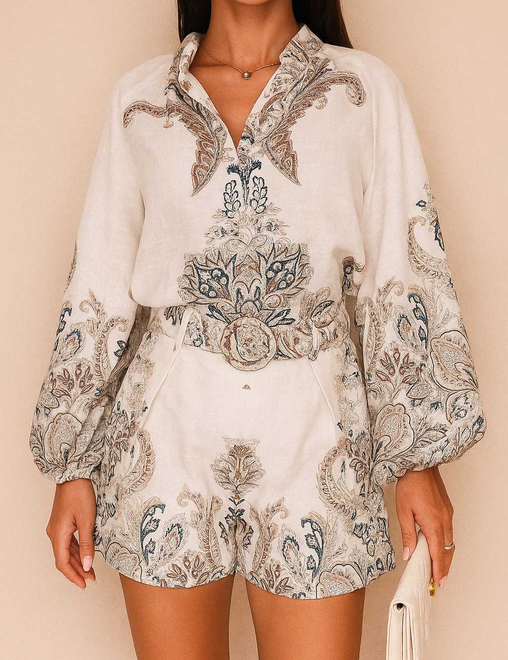 Front view of a women’s grey paisley long-sleeve blouse with split V neckline and billowed sleeves paired with belted shorts featuring slant pockets