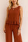 Front detail view of a women’s rust sleeveless ruffle-hem top with scoop neckline paired with wide-leg pants on an elastic waist.