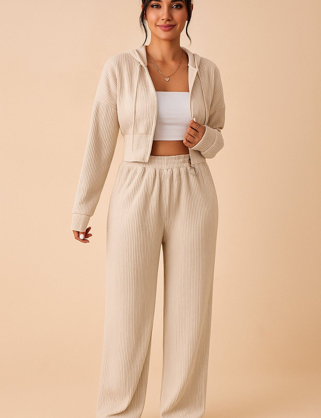 Pastel beige ribbed jumpsuit featuring zip‑up hoodie and elastic waistband, model kneeling.