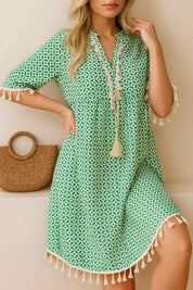 Turquoise geometric-print tassel-trim mini dress with tassel-tie neckline and short sleeves — seated front three-quarter view.